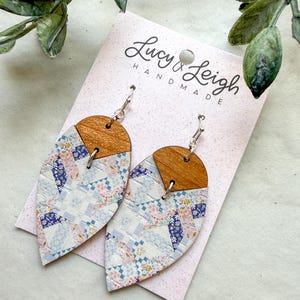 Puede incluir: Pendientes en forma de lágrima con una sección superior de madera natural y una inferior con un estampado floral azul, rosa y blanco inspirado en el patchwork. Están sujetos a una tarjeta blanca con el texto Lucy & Leigh Handmade.