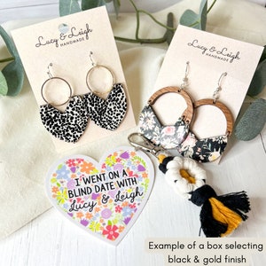 May include: A set of handmade earrings with a black and white animal print design, a floral print design, and a wooden design. The earrings are displayed on a white card with the text "Lucy & Leigh Handmade". There is also a heart-shaped sticker with the text "I went on a blind date with Lucy & Leigh" and a black and gold tassel keychain.