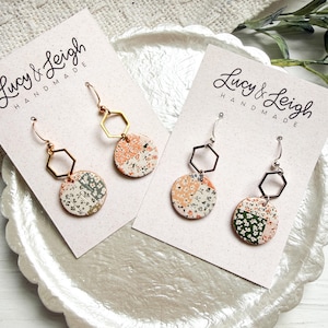 以下が含まれることがあります： 手作りのイヤリングが2組。各イヤリングは、花柄のディスク、幾何学的な形、金属製のフックが特徴です。カードはスカラップトレイにあります。「Lucy & Leigh Handmade」の文字が見えます。