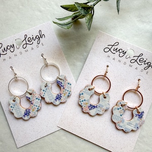 May include: Two pairs of dangle earrings on Lucy & Leigh Handmade display cards. Each earring features a scalloped, U-shaped charm with a blue and white patchwork floral pattern, suspended from a simple circular metal hoop with a hook wire.