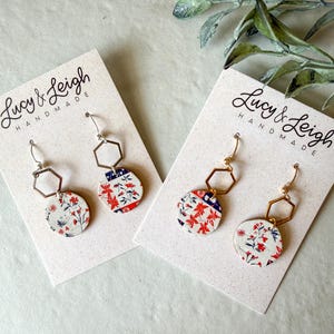 Puede incluir: Dos pares de pendientes colgantes en tarjetas de presentación de Lucy & Leigh Handmade. Cada pendiente cuenta con un conector hexagonal plateado o dorado unido a un colgante circular con un patrón floral en blanco, rojo y azul.