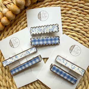May include: Four hair clips with blue and white patterns. Two clips feature a blue and white gingham pattern, and two clips feature a white background with blue floral designs. The clips are on a white background with the text "Lucy & Leigh" in black.