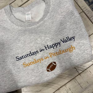 May include: A light gray crewneck sweatshirt featuring embroidered text that reads "Saturdays in Happy Valley" in dark navy blue and "Sundays in Pittsburgh" in gold thread, with a small brown and white football icon centered below the text.