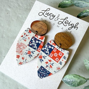 Puede incluir: Pendientes colgantes con postes ovalados de madera oscura unidos a colgantes en forma de escudo. Los colgantes tienen una base blanca con un patrón de retazos de diseños florales rojos y azules. La tarjeta de presentación tiene el texto Lucy & Leigh HANDMADE.