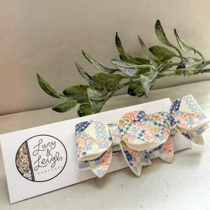 May include: A multi-layered hair bow featuring a patchwork pattern of blue, pink, and white floral and geometric prints. The bow is attached to a white display card with the circular logo text Lucy & Leigh Handmade printed in black ink.