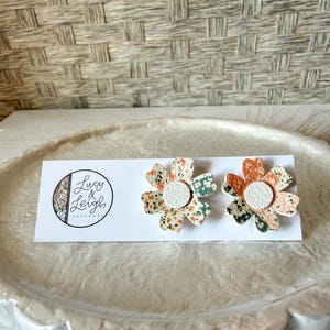 Puede incluir: Pendientes de botón florales con un centro de cuero blanco y un colorido estampado floral en naranja, verde y crema. Los pendientes se muestran en una tarjeta blanca con el logotipo "Lucy & Leigh Handmade".