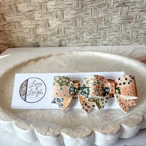 May include: A set of three patterned hair bows with a floral design in peach, green, and navy blue. The bows are attached to a white card with the text "Lucy & Leigh Handmade". The bows are displayed on a white, scalloped tray.