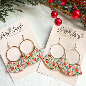 May include: Two pairs of hoop earrings with a festive design. The earrings feature gold-colored hoops and fan-shaped pendants with a floral and nutcracker print in pink, green, and red. The earrings are displayed on a light pink card with the text "Lucy & Leigh HANDMADE".
