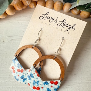 May include: Handmade earrings featuring a wooden half-circle design with a blue and white cherry and bow pattern. The earrings have silver-colored hooks and are displayed on a card that reads "Lucy & Leigh HANDMADE."