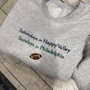 May include: A light grey crewneck sweatshirt featuring embroidered text that reads "Saturdays in Happy Valley" in dark blue and "Sundays in Philadelphia" in dark green, with a small brown and white football icon centered below the text.