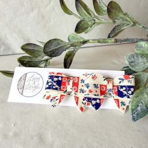 Puede incluir: Un lazo para el cabello con un patrón de retazos de diseños florales y geométricos en rojo, blanco y azul marino. El lazo está sujeto a una tarjeta de presentación blanca con el texto Lucy & Leigh Handmade en un logotipo circular a la izquierda.