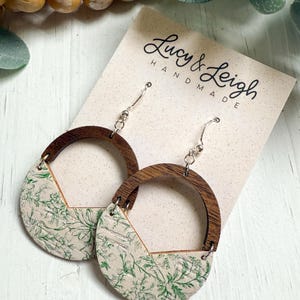 May include: A pair of handmade earrings with a wooden half-circle design. The earrings feature a cream-colored section with a green floral pattern. The earrings are suspended from silver-colored hooks. The tag in the background reads "Lucy & Leigh Handmade".