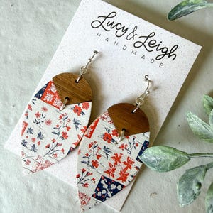 Puede incluir: Un par de pendientes colgantes con una parte superior de madera marrón oscuro en forma de semicírculo unida a una base blanca en forma de hoja con un estampado floral rojo y azul. Los pendientes se presentan en una tarjeta blanca Lucy & Leigh Handmade.