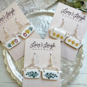 May include: Handmade earrings in the shape of miniature casserole dishes. The earrings are white with various decorative elements, including vegetables, floral patterns, and yellow flowers. The brand name "Lucy & Leigh" is printed on the cards.