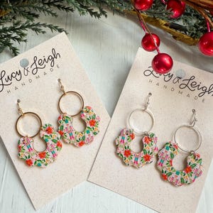 Puede incluir: Dos pares de pendientes hechos a mano con un estampado floral y navideño. Los pendientes presentan aros dorados o plateados y colgantes en forma de flor con detalles rojos, verdes y rosas. Se exhiben en tarjetas de color neutro, con follaje festivo.