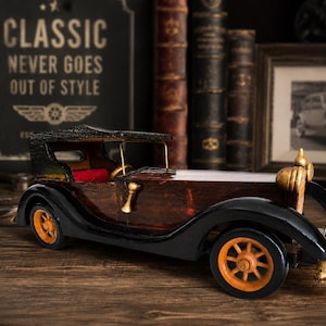 May include: A wooden model car featuring a dark brown body, black fenders, and orange wheels. The vehicle has a black roof, a bright red interior seat, gold-colored headlights, and a gold-colored horn mounted on the side of the driver's door.