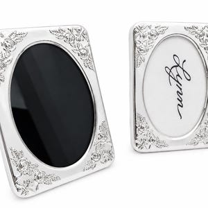 May include: Two rectangular silver-toned picture frames with rounded corners and an oval-shaped central opening. The frames feature embossed floral and leaf detailing at each corner. One frame contains a black insert, while the other displays the name Lynn.