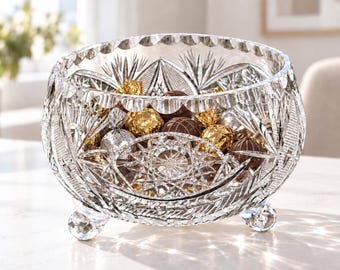 May include: A clear, footed crystal bowl featuring intricate cut-glass patterns, including starburst and fan motifs. The bowl is filled with assorted round chocolates wrapped in gold and silver foil, showcasing a scalloped rim and three small feet.