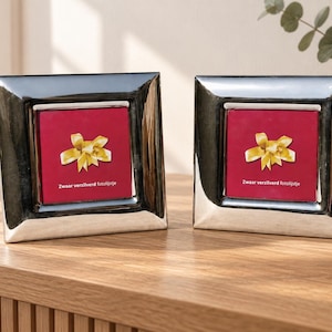 May include: Two square silver-toned picture frames with a wide, polished border. Each frame contains a red insert featuring a yellow ribbon graphic and the text Zwaar verzilverd fotolijstje printed in white at the bottom of the red square area.