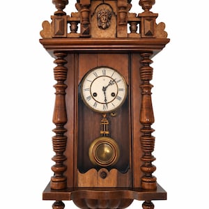 May include: A dark brown wooden wall clock featuring a circular white face with black Roman numerals and ornate hands. The clock is framed by turned wooden pillars and topped with a carved face motif. A brass pendulum hangs below the clock face.