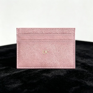 May include: A dusty rose pink leather card holder featuring a textured grain finish and two horizontal card slots. The center of the card holder is embossed with the gold-colored capital letters DC, centered in a clean, sans-serif font.