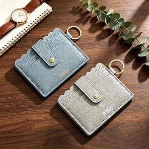 May include: Two rectangular card holders with scalloped edges and gold-tone key rings. One is light blue with the name Janie embossed in gold, and the other is light gray with the name Riley embossed in white. Both feature a snap-button strap.