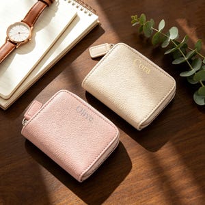 Puede incluir: Dos carteras pequeñas rectangulares con cremallera y acabado texturizado. Una cartera es de color rosa claro con el nombre Olive grabado en el frente, y la otra es de color crema con el nombre Cora grabado en el frente. Ambas tienen tirador.