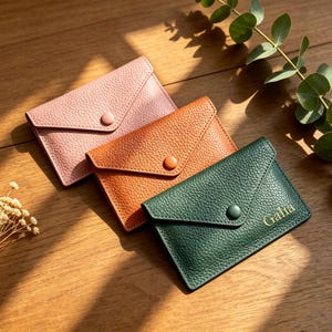 May include: Three envelope-style card holders with a pebbled texture, each featuring a matching snap button closure. The holders are colored light pink, orange, and dark green, with the name Galia embossed in gold on the dark green holder.