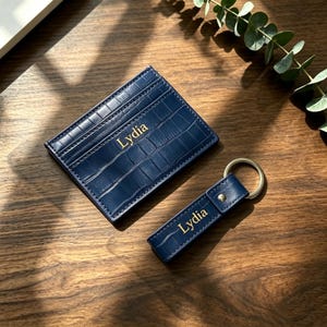 Peut inclure: Un porte-cartes bleu marine et un porte-clés rectangulaire assorti, tous deux dotés d'une texture gaufrée façon crocodile et du nom Lydia inscrit en lettres dorées. Le porte-clés comprend un anneau métallique et un rivet doré.