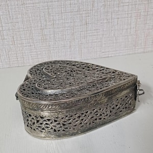 May include: A silver-colored, heart-shaped box with intricate openwork designs. The box has a hinged lid and a clasp closure. The metalwork features floral and geometric patterns, creating a delicate and ornate appearance. The box is likely used for jewelry or small keepsakes.