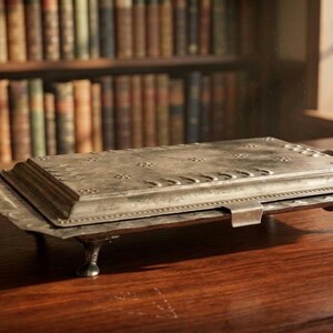 Puede incluir: Una caja rectangular de metal plateado con tapa abatible, que presenta patrones en relieve de flores y lunas crecientes. La caja se apoya sobre cuatro patas pequeñas y curvas, y tiene una base ancha con un borde decorativo festoneado.