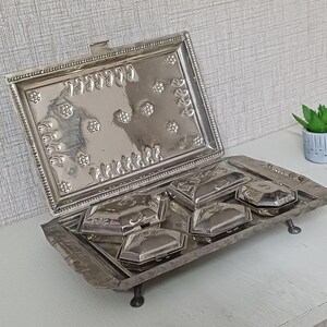 Puede incluir: Una bandeja de servicio rectangular de metal plateado con tapa abatible decorada con patrones florales y de volutas en relieve. La base contiene cuatro recipientes hexagonales extraíbles con tapa, superficies pulidas y patas.