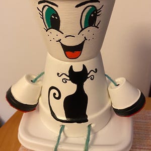 Puede incluir: Una maceta pintada a mano y caprichosa con forma de personaje sonriente. La maceta blanca presenta un diseño de gato negro, ojos verdes y boca roja. La maceta tiene detalles en blanco y negro y está sobre un recipiente blanco.
