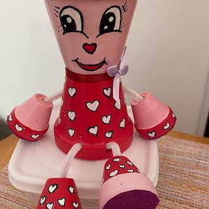 Peut inclure: Une poupée en pot de terre cuite peinte à la main. La poupée a un corps rouge orné de cœurs blancs, des bras et des jambes roses, et un visage souriant. Un nœud violet orne le cou. La poupée est assise sur un récipient blanc.