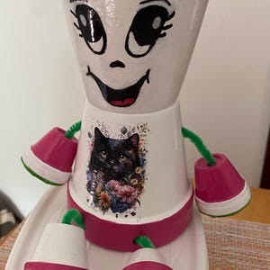 Peut inclure: Pot de fleur fantaisiste peint à la main, en forme de personnage souriant. Le pot blanc présente un motif de chat noir entouré de fleurs. Le pot a des accents roses, des bras en cure-pipe verts et des pieds assortis.