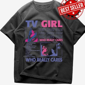 May include: A charcoal gray t-shirt featuring the text TV GIRL and WHO REALLY CARES in blue and pink. The graphic includes a tracklist of ten songs and two framed illustrations of figures in blue and pink tones. A red BEST SELLER badge is visible.