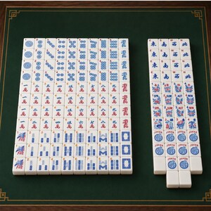 May include: A complete Mahjong set with white tiles featuring blue and red symbols and characters. The tiles are arranged on a green playing surface with a dark wood border. Includes tiles with numbers, suits, and Jokers.