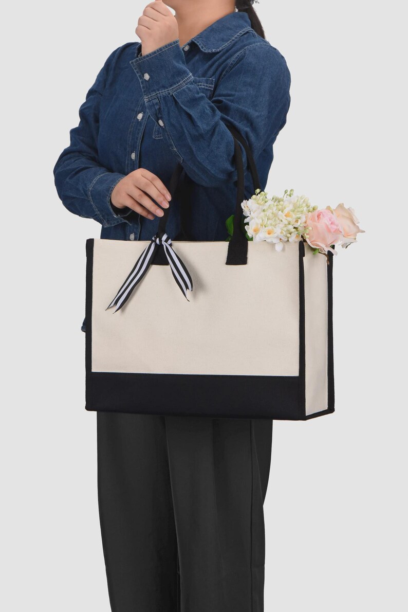 Peut inclure: Un sac fourre-tout rectangulaire en toile avec un corps couleur cr&egrave;me et une base noire unie. Le sac pr&eacute;sente des anses en tissu noir et un ruban d&eacute;coratif &agrave; rayures noires et blanches nou&eacute; &agrave; une anse. L'int&eacute;rieur contient un bouquet de fleurs roses et blanches.