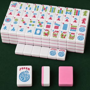 May include: A complete Mahjong set with white tiles featuring various symbols, numbers, and Chinese characters in red, blue, and green. Includes joker tiles and pink and white accessories. The tiles are arranged on a green surface.