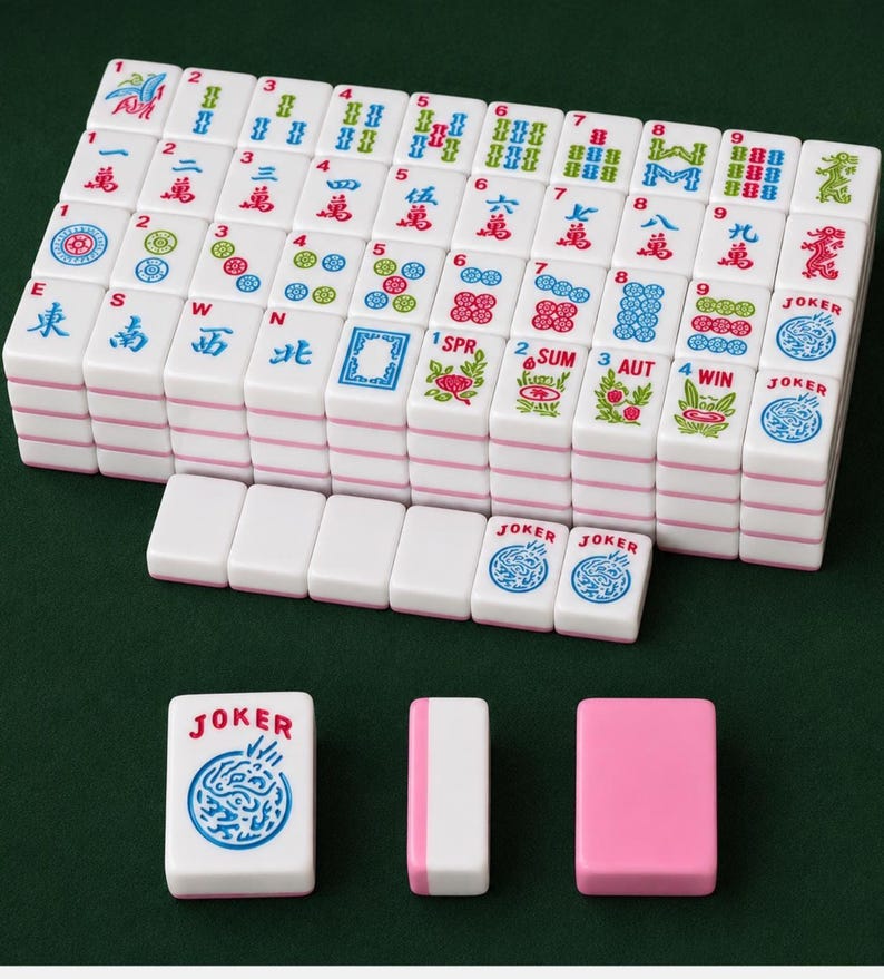 May include: A complete Mahjong set with white tiles featuring various symbols, numbers, and Chinese characters in red, blue, and green. Includes joker tiles and pink and white accessories. The tiles are arranged on a green surface.