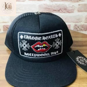 Puede incluir: Gorra de camionero negra con parte trasera de malla y visera plana. La parte delantera presenta un parche rectangular con el texto "CHROME HEARTS HOLLYWOOD, USA" y un gráfico de labios rojos con un diente dorado. Pequeño emblema plateado en la parte superior.