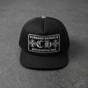 Puede incluir: Gorra de camionero negra con parte trasera de malla y visera curva. El panel frontal presenta un parche bordado blanco con el texto "CHROME HEARTS HOLLYWOOD, USA". Un botón de metal plateado está en la parte superior.