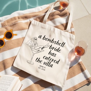 May include: A cream-colored canvas tote bag featuring a black line drawing of a bride sitting in a martini glass. The text reads: a bombshell bride has entered the villa, MIAMI, FL, EST. 2026. The bag has two shoulder straps and a rectangular shape.