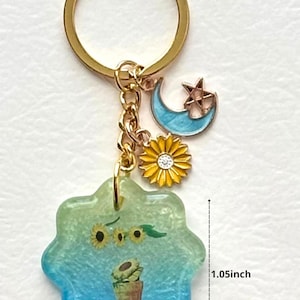 May include: A gold-colored keychain with a blue and green flower-shaped charm, measuring 1.05 inches across. The charm features a sunflower design and a small potted sunflower. Additional charms include a yellow sunflower, a blue crescent moon, and a star.