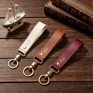 May include: Three leather keychains in white, brown, and burgundy, each featuring a gold-tone metal clasp and ring. The white keychain is embossed with the name Kevin, the brown with Peter, and the burgundy with Scott, all in gold lettering.