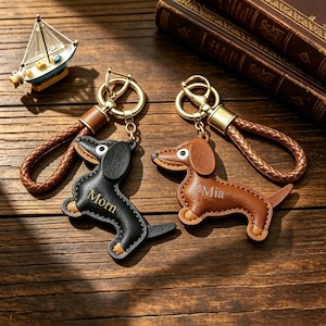 May include: Two dachshund-shaped keychains with braided leather loops and gold-tone metal rings. One keychain is black with the name Morn embossed in gold, and the other is brown with the name Mia embossed in gold. Both feature stitched edges.