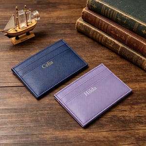 May include: Two rectangular card holders with a textured finish. One is dark blue with the name Celia embossed in gold, and the other is light purple with the name Hilda embossed in silver. Both feature multiple slots for organizing credit cards.