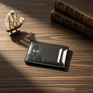 May include: A black rectangular card holder featuring an embossed graphic of a masked superhero character on the left side. The accessory has white stitching along the edges and a diagonal slot on the right side containing a white card insert.