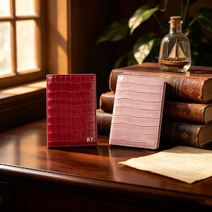 May include: Two leather journals with an embossed crocodile texture. One is deep red, the other is a soft pink. Each journal has gold initials. The journals are on a wooden surface, with antique books and a decorative glass bottle in the background.