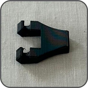Peut inclure: Un objet noir angulaire avec une section incurvée et deux protubérances rectangulaires. L'objet a une surface texturée avec des reflets bleus et verts. Il est posé sur un fond gris clair texturé.